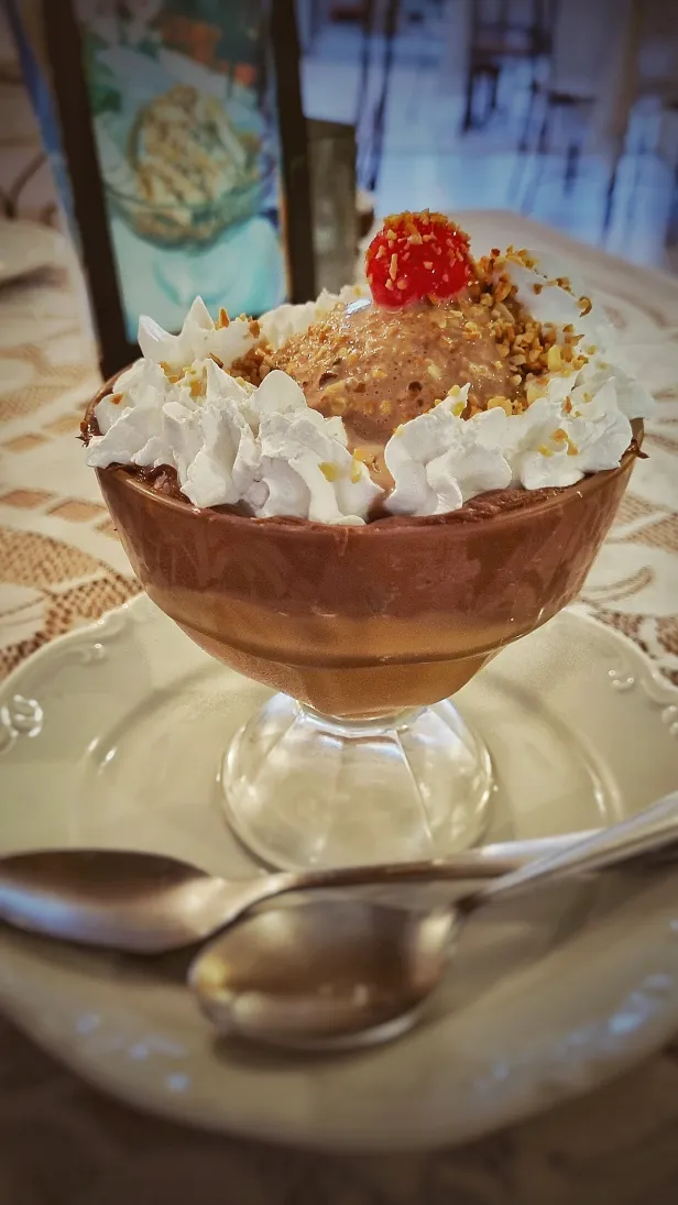 Taça de sorvete de chocolate coberto com chantilly, farelo de amendoim e uma cereja. No pires, duas colheres.