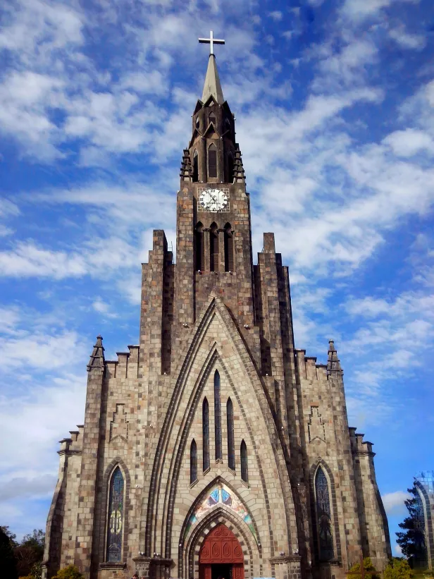 Catedral de grandes proporções com portas em madeira, mas construída toda em pedras cinzentas. Sua alta torre ostenta um relógio branco e uma cruz no topo.