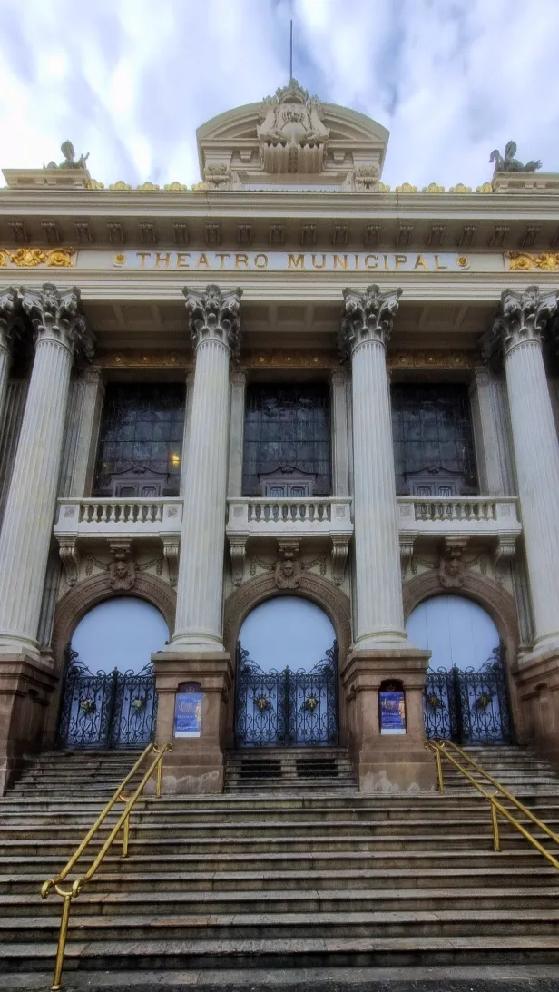 Escadaria em pedra até três portões em arco, com colunas decoradas que passam o segundo pavimento de grandes janelas até o beiral decorado. Na fachada, lê-se Theatro Municipal em dourado.