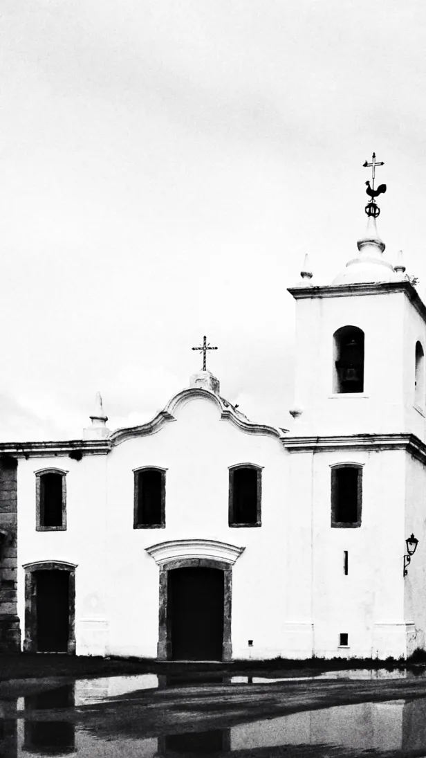 Igreja colonial em preto e branco, com alto contraste, refletida em poças d'água no chão. No alto da torre, a silhueta de um galo e de uma cruz.