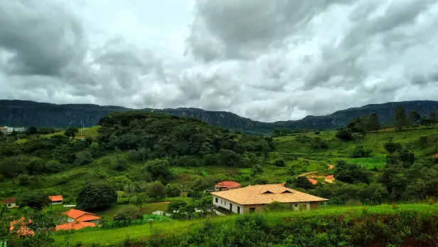 Área verde bem arborizada, com casas algumas coloniais. Ao fundo, uma formação montanhosa cobre todo o horizonte sob um céu nublado.