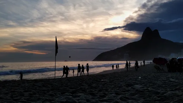 Silhueta de pessoas jogando uma bola ao ar, próximo às ondas da praia no fim de tarde. Em foco, uma bandeira fincada na areia. Ao fundo, uma montanha com dois grandes picos.