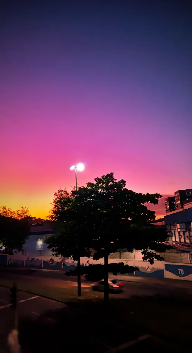 Céu urbano em degradê de azul escuro, violeta e laranja. A silhueta de uma árvore esconde parte de um poste com luzes acesas. Um carro passa pela rua.