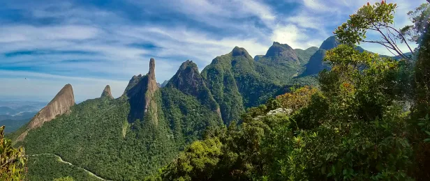 Formação montanhosa e arborizada, com ao menos sete picos rochosos bem proeminentes, vista ao nível da foto. Céu azul com algumas nuvens.