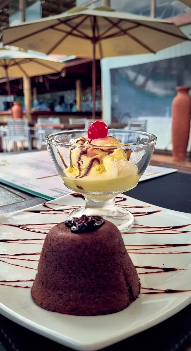 Pequeno bolo de chocolate em um prato decorado com calda, junto de uma taça de sorvete de creme com cereja. Desfocados ao fundo, mesas com ombrelones.
