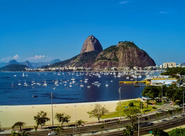 Famoso morro com duas formações rochosas em uma enseada repleta de barcos em um dia de Sol e céu limpo.