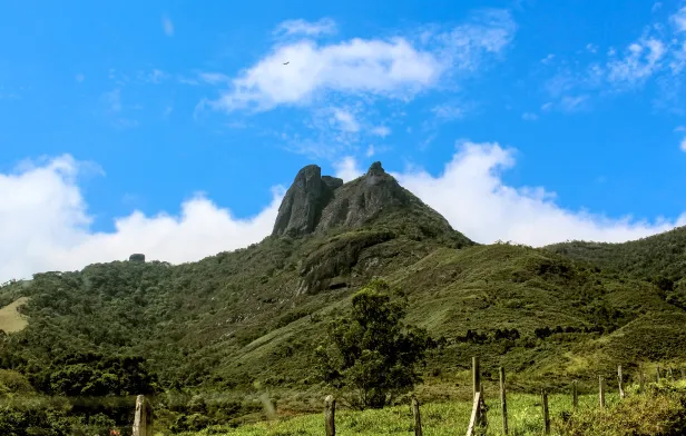 Área de aclive com vegetação rasteira, cercas de arame e uma árvore ao centro. Distante, ao fundo, uma montanha com dois picos rochosos. Céu azul com poucas nuvens.