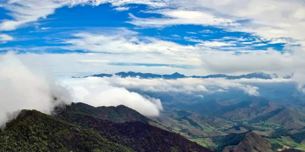 Nuvens vistas de cima com parte do céu em um azul vívido. Região montanhosa de verde abundante, com picos distantes no horizonte.