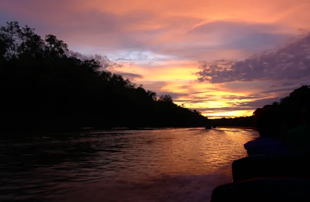 Rio cercado pela silhueta de florestas altas com o nascer do Sol ao fundo, com tons de azul, lilás e laranja refletidos nas pequenas ondulações formadas pela embarcação, completamente sombreada.