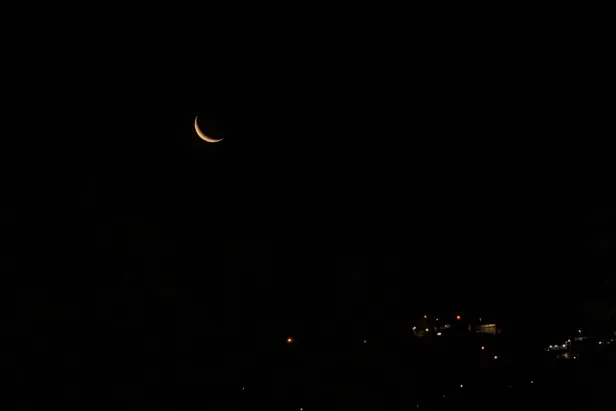 Lua crescente de coloração alaranjada e alguma iluminação urbana na parte inferior.