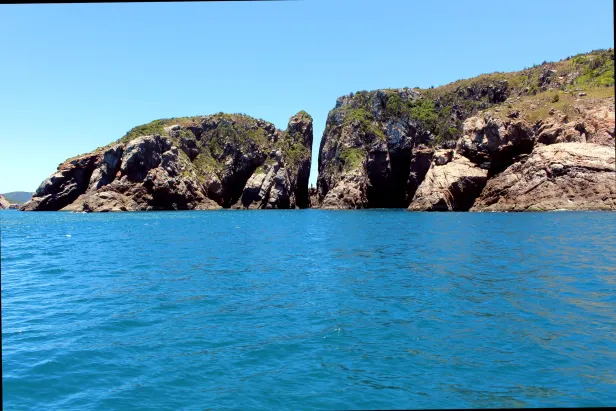 Vista do mar para uma formação rochosa dividida por uma fenda, onde se nota algo em pé, mas indistinguível. A formação à esquerda da fenda lembra a de uma tartaruga de perfil.
