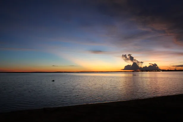 Pôr do Sol com poucas nuvens ao fundo, sobre um corpo d'água com leves ondulações até a orla.