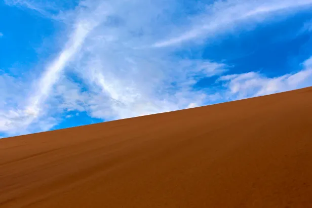 Duna de areia de tom marrom vívido cortando a tela na diagonal, com o céu azul com poucas nuvens brancas.