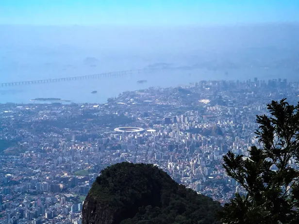 Visão de cima de área urbana densa, com um estádio de futebol ao centro. Ao fundo, uma ponte atravessa o horizonte de um lado ao outro.